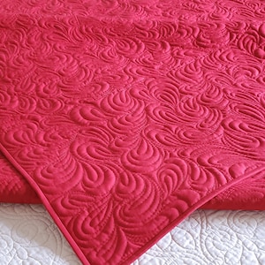May include: A red quilted blanket with a swirling pattern. The blanket is folded in half and is lying on a white surface.