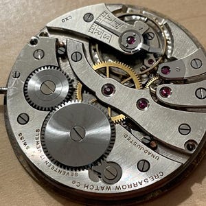 May include: Close-up of a silver watch movement with the text "Cresarrow Watch Co. Seventeen Jewels Swiss Unadjusted" on the outer edge. The movement has a gold-colored winding mechanism and several gears.