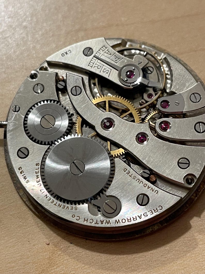 May include: Close-up of a silver watch movement with the text "Cresarrow Watch Co. Seventeen Jewels Swiss Unadjusted" on the outer edge. The movement has a gold-colored winding mechanism and several gears.