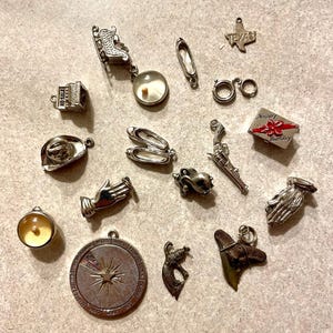 May include: A collection of silver charm pendants, including a compass, a shark tooth, a cowboy hat, a pair of ballet shoes, a gun, a building, a hand, a pair of praying hands, a Christmas sleigh, a Texas state outline, a birthday present, and a mermaid.