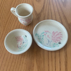 May include: A set of three ceramic dishes with a rooster design. The dishes are white with pink, green, and blue accents. The set includes a small bowl, a larger bowl, and a plate.