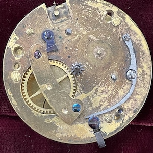 Op de afbeelding: Close-up van een messing uurwerk met tandwielen en een opwindslinger. Het uurwerk is gedemonteerd en toont de ingewikkelde werking van het horloge.