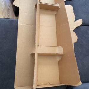 Op de afbeelding: Een kartonnen doos met twee uitgesneden secties, waarschijnlijk voor het verpakken van een product. De doos is bruin en heeft een eenvoudig ontwerp.