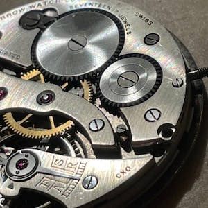 May include: Close-up of a silver watch movement with the text "Arrow Watch Co. Seventeen 17 Jewels Swiss Adjusted" and "FAR" and "CXO" visible.