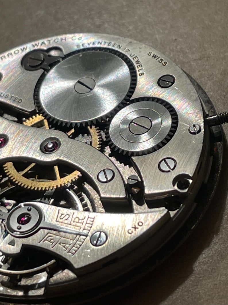 May include: Close-up of a silver watch movement with the text "Arrow Watch Co. Seventeen 17 Jewels Swiss Adjusted" and "FAR" and "CXO" visible.