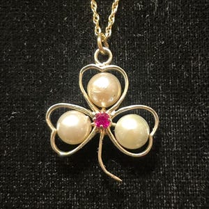 May include: A gold chain necklace with a pendant shaped like a four-leaf clover. The clover has three white pearls and a red gemstone in the center.