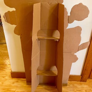 Op de afbeelding: Een kartonnen stand-up cutout van een cartoonfiguur. De cutout is bruin en heeft twee planken. De cutout staat op een houten vloer.