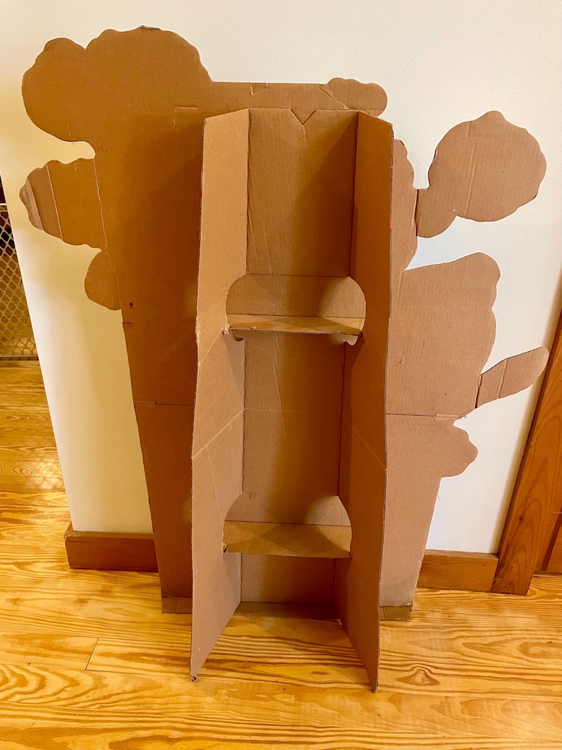 Op de afbeelding: Een kartonnen stand-up cutout van een cartoonfiguur. De cutout is bruin en heeft twee planken. De cutout staat op een houten vloer.