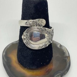 May include: A silver spoon ring with ornate floral detailing. The ring is wrapped around a black velvet ring holder, showcasing the intricate design. The spoon's bowl and handle are visible, reflecting light.