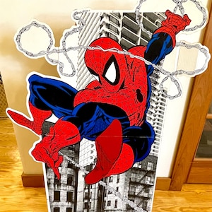 Op de afbeelding: Een kartonnen uitsparing van Spider-Man die door een stadsbeeld zwaait. Hij draagt zijn klassieke rode en blauwe pak met een spinnenlogo op zijn borst. De achtergrond is een zwart-wit beeld van gebouwen.