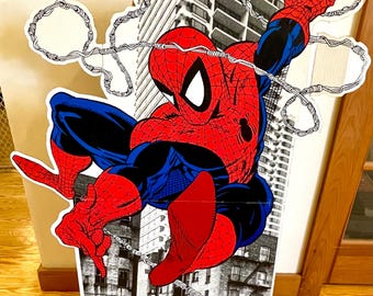 May include: A cardboard cutout of Spider-Man swinging through a city skyline. He is wearing his classic red and blue suit with a spider logo on his chest. The background is a black and white image of buildings.