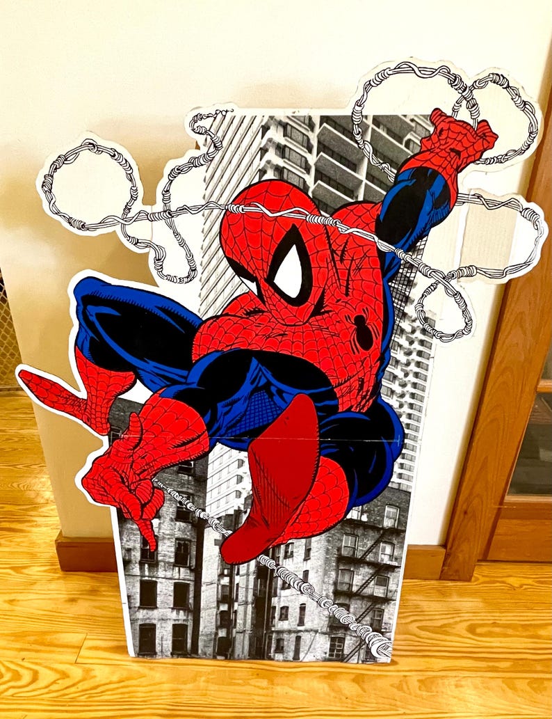 Op de afbeelding: Een kartonnen uitsparing van Spider-Man die door een stadsbeeld zwaait. Hij draagt zijn klassieke rode en blauwe pak met een spinnenlogo op zijn borst. De achtergrond is een zwart-wit beeld van gebouwen.