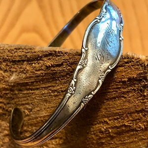 May include: A silver spoon bracelet with floral and scroll detailing. The spoon's bowl is engraved with the initials "M.L.D." The bracelet is bent into a cuff shape and rests on a piece of wood.