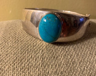Op de afbeelding: Een zilveren armband met een turquoise cabochon-steen in het midden.