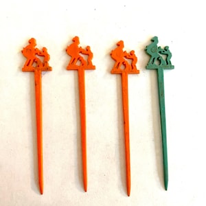 May include: Four vintage cocktail picks. Three are orange, and one is green. Each pick features a decorative silhouette of a person with two children. The picks are made of plastic and have a pointed end.