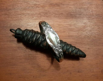 Op de afbeelding: Een zilveren lepelring met bloemendetails rust op een donker, getextureerd, dennenappelvormig object. Het gepolijste oppervlak van de ring contrasteert met de ruwe textuur van de donkerbruine dennenappel. De achtergrond is een warm houten oppervlak.