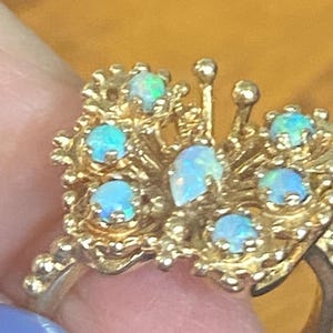 Op de afbeelding: Een gouden ring met een cluster van opaalstenen gezet in een delicaat, ingewikkeld ontwerp. De ring heeft een verscheidenheid aan opaalvormen en -maten, waardoor een uniek en opvallend sieraad ontstaat.