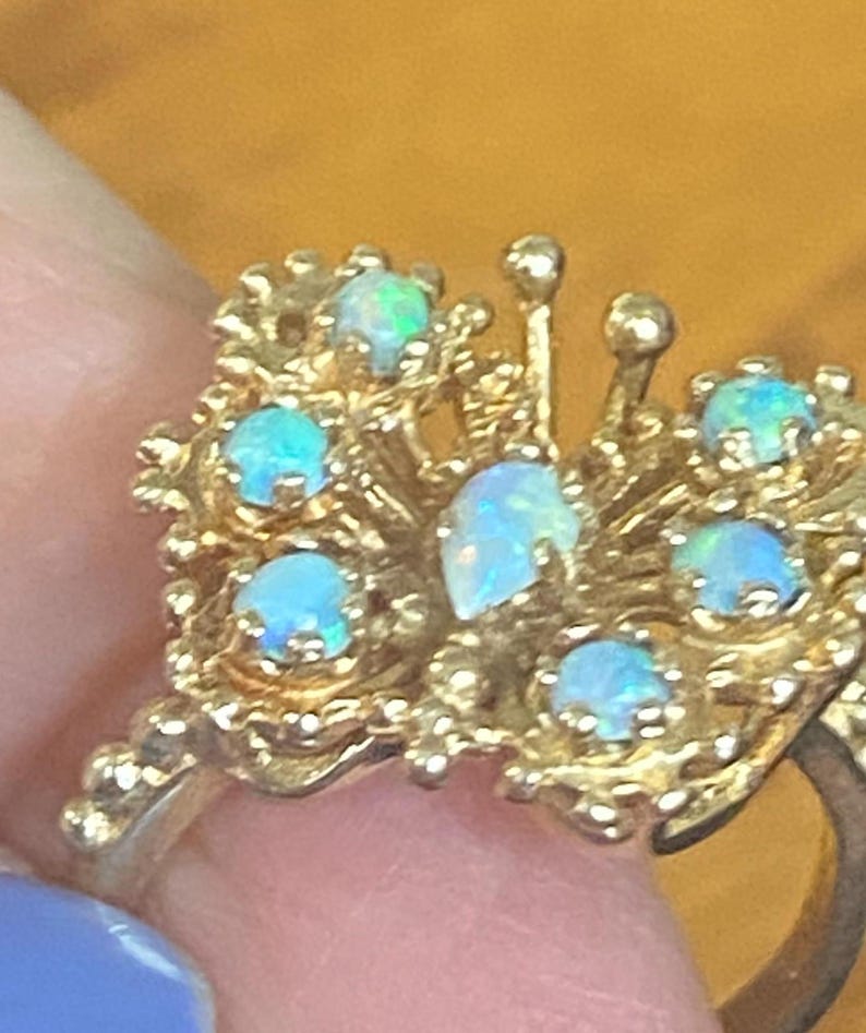 Op de afbeelding: Een gouden ring met een cluster van opaalstenen gezet in een delicaat, ingewikkeld ontwerp. De ring heeft een verscheidenheid aan opaalvormen en -maten, waardoor een uniek en opvallend sieraad ontstaat.