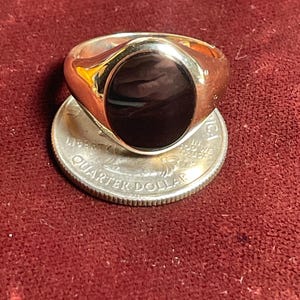 May include: A gold-toned signet ring with a large, oval, black stone set in the center. The ring is resting on a US quarter coin, showcasing its size and design against a red background.