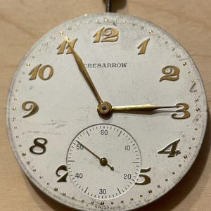 May include: A white watch face with gold-colored numerals and hands. The watch face has the text "CRESARROW" and "SWISS" printed on it. The watch face is surrounded by a gold-colored ring with small dots.