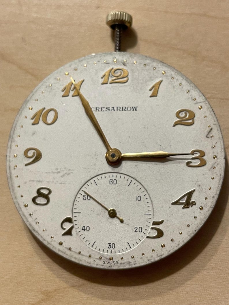 May include: A white watch face with gold-colored numerals and hands. The watch face has the text "CRESARROW" and "SWISS" printed on it. The watch face is surrounded by a gold-colored ring with small dots.