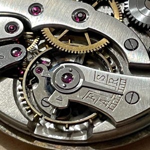 May include: Close-up of a silver watch movement with intricate gears and a red jewel. The movement is labeled "ETA" and "SAR".