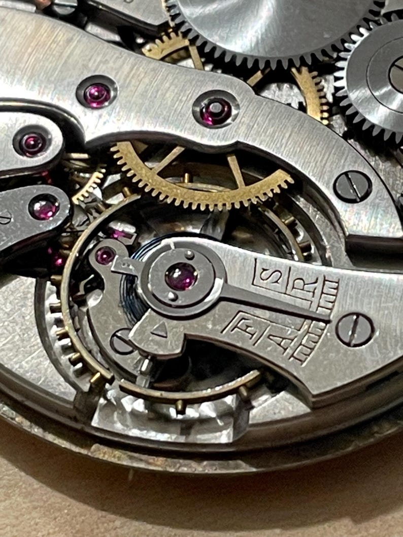 May include: Close-up of a silver watch movement with intricate gears and a red jewel. The movement is labeled "ETA" and "SAR".