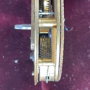 Op de afbeelding: Close-up van een goudkleurig uurwerkmechanisme met ingewikkelde tandwielen en een ketting. Het mechanisme is geplaatst tegen een donkerrode achtergrond.