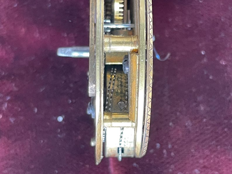 Op de afbeelding: Close-up van een goudkleurig uurwerkmechanisme met ingewikkelde tandwielen en een ketting. Het mechanisme is geplaatst tegen een donkerrode achtergrond.