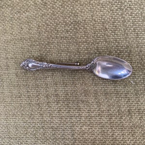 May include: A silver spoon with an ornate handle, the spoon is lying on a textured green surface.