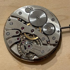 May include: Close-up of a silver watch movement with the text "Cres Sarrow Watch Co. Seventeen Jewels Swiss Unadjusted" and "CXO" visible. The movement has a gold-colored winding stem and several gears.