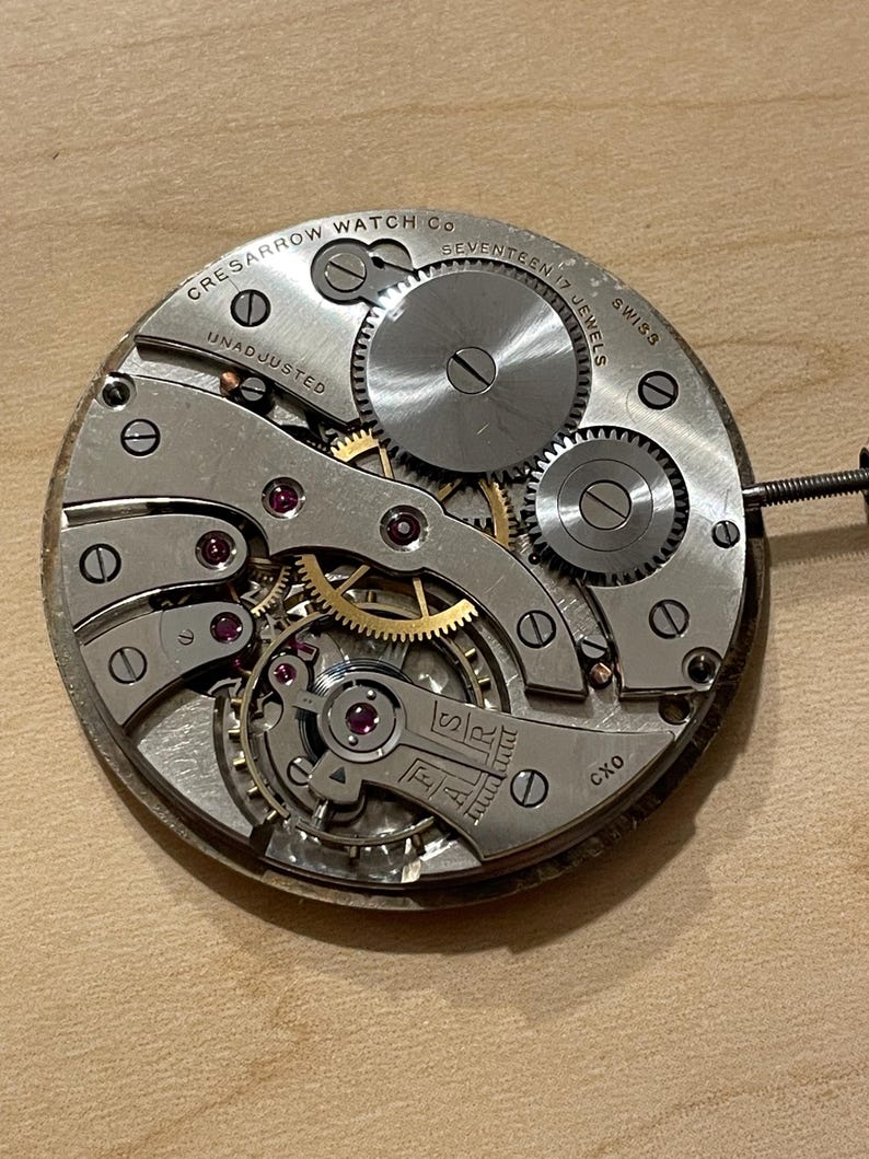 May include: Close-up of a silver watch movement with the text "Cres Sarrow Watch Co. Seventeen Jewels Swiss Unadjusted" and "CXO" visible. The movement has a gold-colored winding stem and several gears.