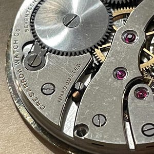 May include: Close-up of a silver watch movement with the text "Crescent Watch Co. Seventeen Jewels Unadjusted" and "Swiss" visible. The movement has a series of gears and a small screw.