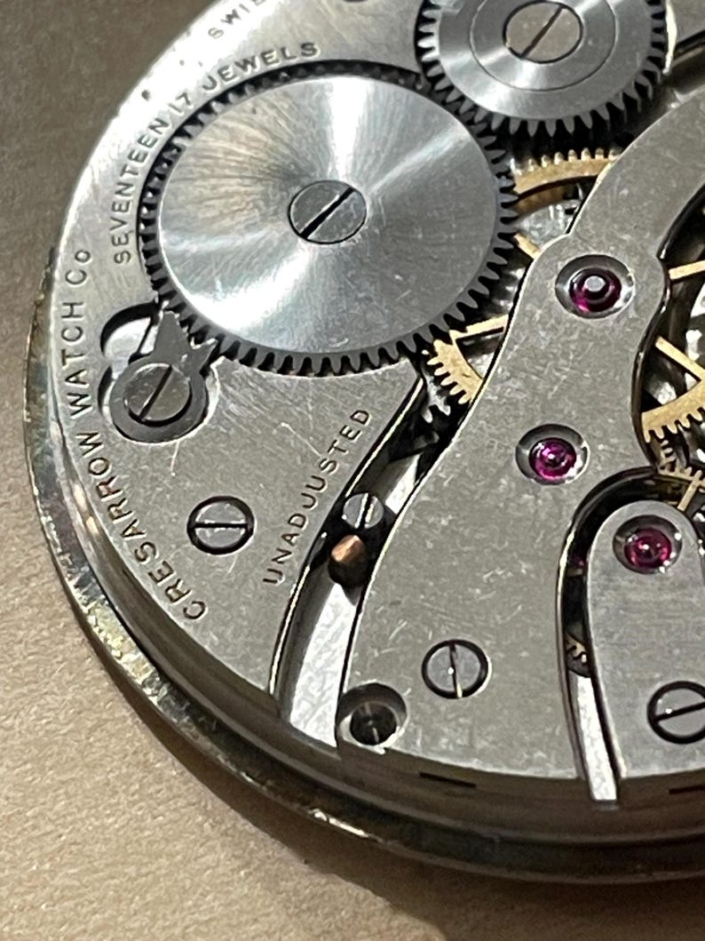 May include: Close-up of a silver watch movement with the text "Crescent Watch Co. Seventeen Jewels Unadjusted" and "Swiss" visible. The movement has a series of gears and a small screw.