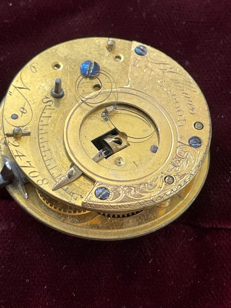 Op de afbeelding: Close-up van een gouden zakhorloge-uurwerk met ingewikkelde gravures en een blauwe stalen schroef. De wijzerplaat van het horloge is zichtbaar met Romeinse cijfers en de tekst "Thomas Moore London".