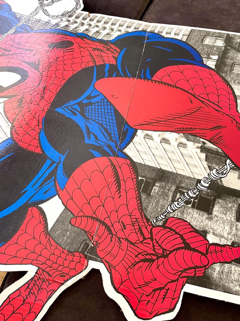 Op de afbeelding: Een rood en blauw Spiderman-actiefiguur met een web shooter in zijn hand, zwaaiend aan een weblijn tegen een stadsachtergrond.