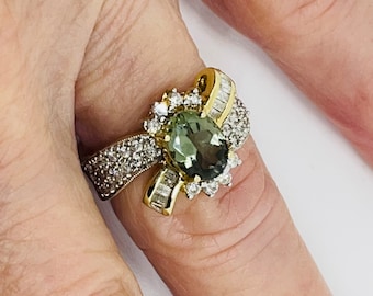 Op de afbeelding: Een gouden ring met een groene ovale edelsteen omringd door kleinere diamanten. De ring heeft een boogvormig ontwerp met een rij baguette-geslepen diamanten aan één kant.
