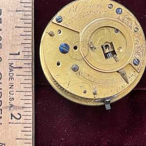 Op de afbeelding: Close-up van een goudkleurig zakhorlogemechanisme met de inscriptie "B. Crower Liverpool" op de wijzerplaat. Het horloge heeft een opwindsleutel en een kleine wijzer.