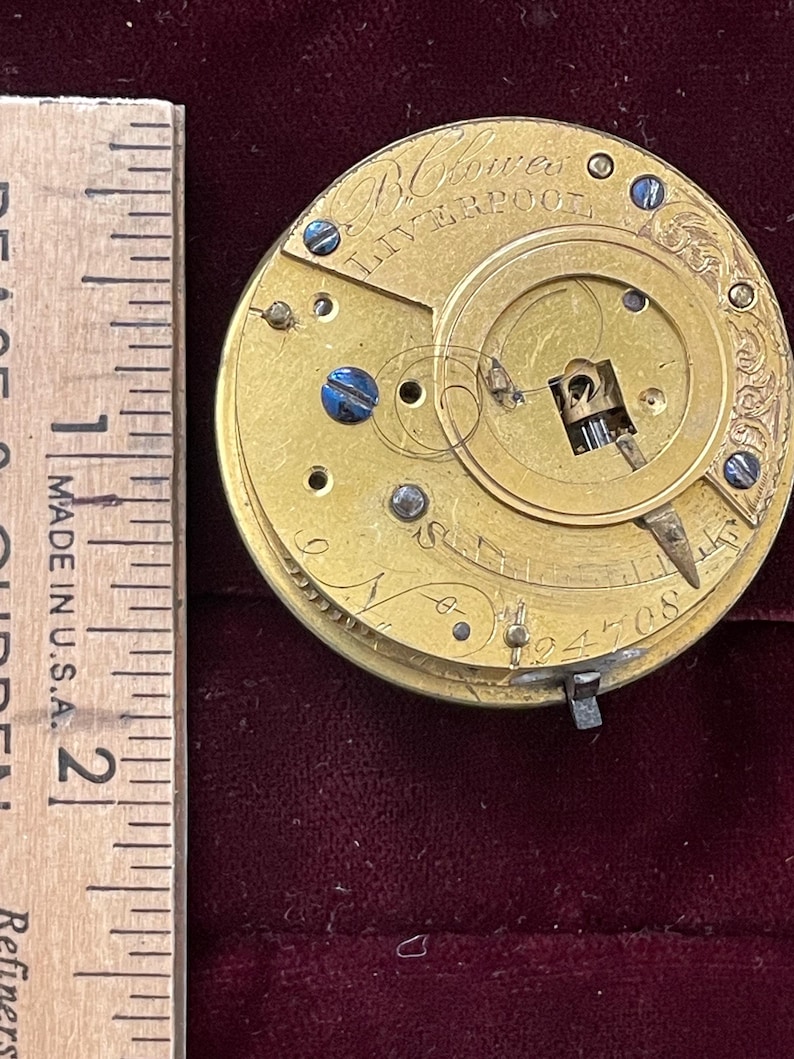 Op de afbeelding: Close-up van een goudkleurig zakhorlogemechanisme met de inscriptie "B. Crower Liverpool" op de wijzerplaat. Het horloge heeft een opwindsleutel en een kleine wijzer.
