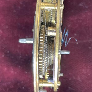 Op de afbeelding: Close-up zijwaartse weergave van een goudkleurig uurwerkmechanisme met een opgewonden veer. Het mechanisme is gemaakt van metalen tandwielen en is zichtbaar door een transparante glazen deksel.