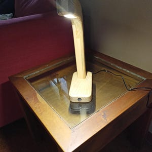 May include: A wooden desk lamp with a rectangular base and a curved arm supporting a rectangular light fixture. The lamp is made of light-colored wood and is turned on, emitting a warm glow. The lamp is on a wooden table.