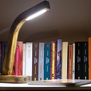 Puede incluir: Una lámpara de escritorio de madera con un brazo curvo ilumina una estantería de libros. La lámpara tiene un acabado de madera natural y una luz LED brillante. Una colección de libros coloridos está dispuesta en la estantería, con un libro plano en primer plano.