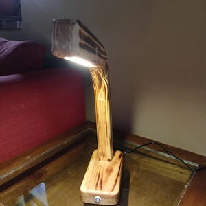 May include: A wooden desk lamp with a rectangular base and a curved arm supporting a rectangular light. The wood has a natural grain and a burnt finish. The lamp is turned on, emitting a bright light.