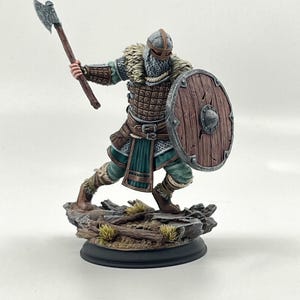 May include: A detailed figurine of a Viking warrior in battle stance, holding an axe and shield. The figure wears a helmet, chainmail, and leather armor, with a fur collar. The shield is round and wooden. The base is a rocky terrain.
