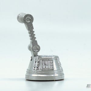 May include: A silver-colored, futuristic-looking desk accessory. It has a cylindrical base with detailed panels and a curved arm ending in a circular head. The arm has a textured, ridged design. The object appears to be made of metal.