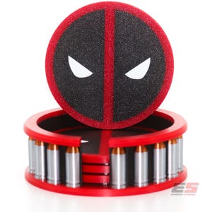 May include: A red and black Deadpool-themed coaster set. The coaster features the character's mask design, with a black background, red border, and white eyes. The base is red and holds several silver bullet-shaped objects.