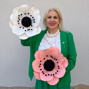 May include: Two large paper flowers, one white and one peach, with black centers and dotted details. The flowers are held up against a woman wearing a green blazer, white lace top, and pearl jewelry. The flowers appear to be handmade.