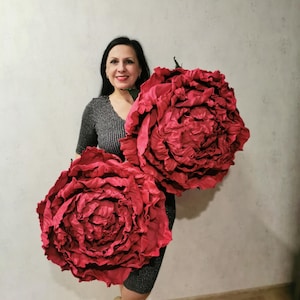 May include: Two large, red, fabric peonies. The peonies are held by a person in front of a white wall.