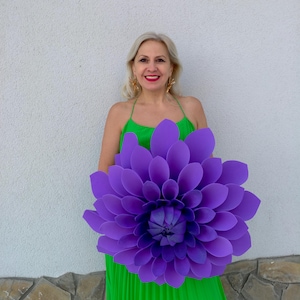 May include: A large, vibrant purple paper flower with layered petals. The flower is held in front of a person wearing a green dress. The flower's colour contrasts with the green dress and the neutral background.