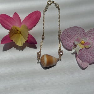 May include: A gold chain necklace with a large, light brown and white shell pendant. The necklace also features small pearls and is displayed with pink and yellow orchids.